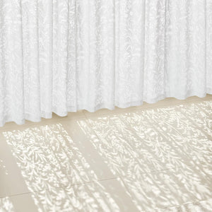 White sheer curtains with a delicate botanical pattern, filtering sunlight onto a light tiled floor.