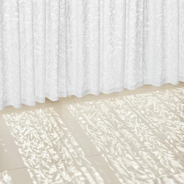 White sheer curtains with a delicate botanical pattern, filtering sunlight onto a light tiled floor.