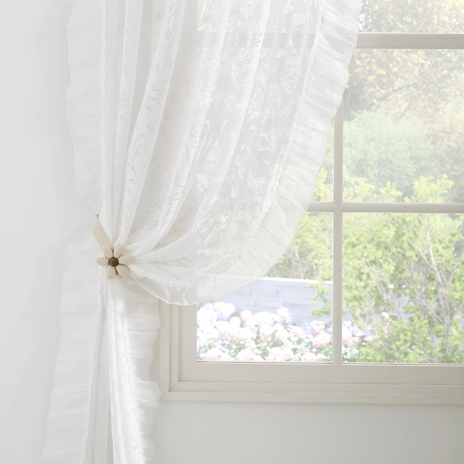 White sheer floral ruffled curtain with cream flower tie-back filters light from sunlit window.