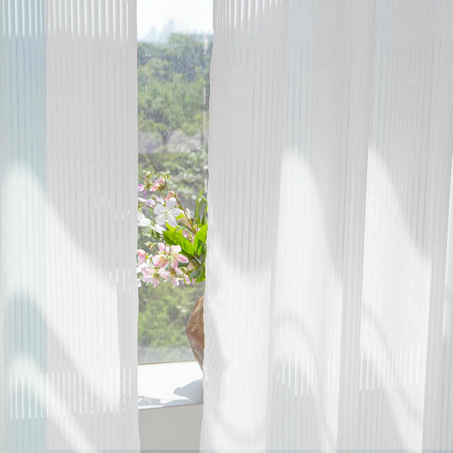 White sheer pleated curtains filtering sunlight by a window, with a soft view of outdoor greenery and flowers.