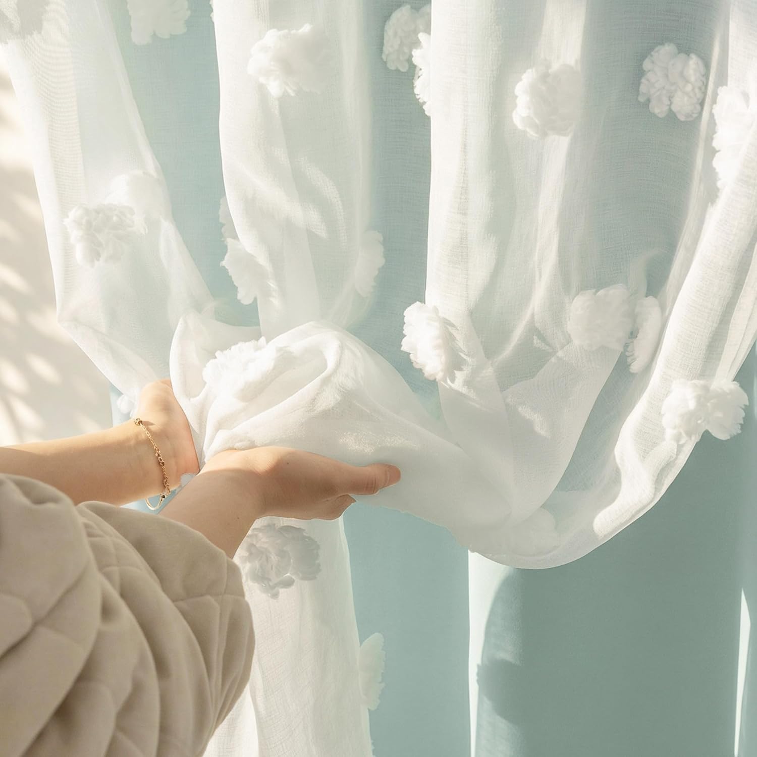 White sheer curtain with fluffy pom-poms, gently held by hands. Sunlight filters through the delicate fabric.