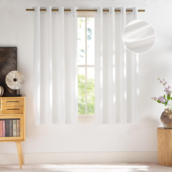 White textured semi-sheer grommet top window curtains hanging on a brass rod in a sunny room.
